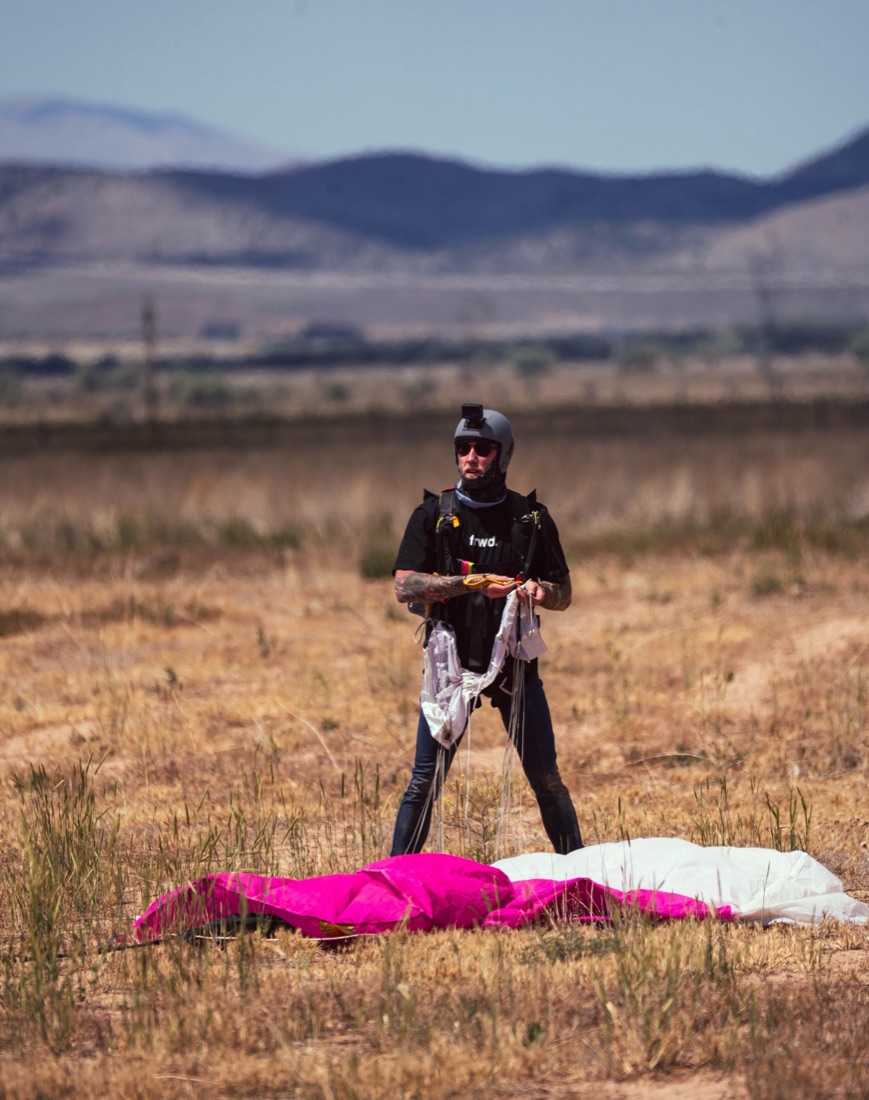 Brock gathering his parachute lines after landing, pink canopy in the grass